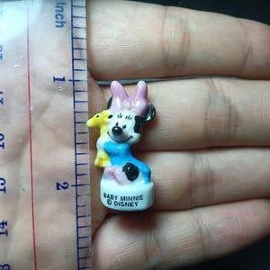 May include: A small Baby Minnie Mouse figurine, approximately 4.4 cm tall, with a pink bow, black ears, and a blue dress. The figurine is holding a yellow plush toy. The base reads "BABY MINNIE DISNEY".