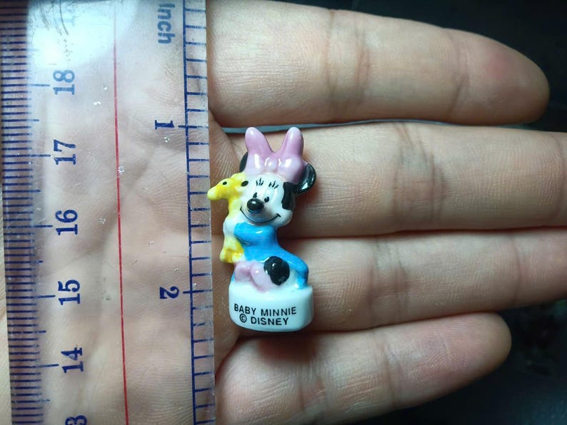 May include: A small Baby Minnie Mouse figurine, approximately 4.4 cm tall, with a pink bow, black ears, and a blue dress. The figurine is holding a yellow plush toy. The base reads "BABY MINNIE DISNEY".
