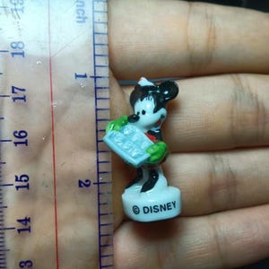 May include: A small Disney Mickey Mouse figurine, approximately 4.4 cm tall, is held in a hand next to a ruler. The figurine is white, black, red, and green, and is holding a blue object. The base of the figurine has the word "DISNEY".