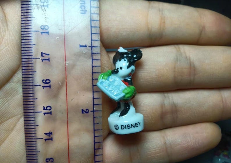 May include: A small Disney Mickey Mouse figurine, approximately 4.4 cm tall, is held in a hand next to a ruler. The figurine is white, black, red, and green, and is holding a blue object. The base of the figurine has the word "DISNEY".