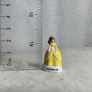 May include: A small, decorative figurine of a woman in a yellow gown with white trim. The figurine is approximately 1.5 inches tall and has the word "CENDRILLON" printed on the base. A ruler is next to the figurine.