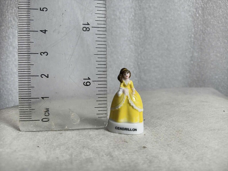 May include: A small, decorative figurine of a woman in a yellow gown with white trim. The figurine is approximately 1.5 inches tall and has the word "CENDRILLON" printed on the base. A ruler is next to the figurine.