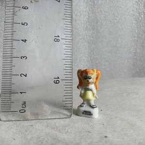 May include: A small ceramic figurine of a girl with orange hair in pigtails, wearing a yellow dress and white top. The figurine is approximately 1 inch tall and stands on a white base with the text "PISTOL DISNEY".