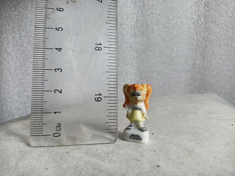 May include: A small ceramic figurine of a girl with orange hair in pigtails, wearing a yellow dress and white top. The figurine is approximately 1 inch tall and stands on a white base with the text "PISTOL DISNEY".