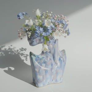May include: A blue ceramic vase shaped like a torso, holding a floral arrangement of white, blue, and pink flowers. The vase has a marbled design in blue and purple hues. The flowers include small blue and larger white flowers.