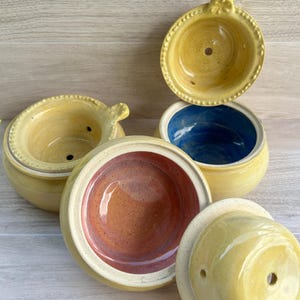 Könnte beinhalten: Eine Sammlung von vier Keramikschalen mit Deckeln, die gelbe Außenseiten und verschiedene Innenfarben wie Blau, Rosa und Gelb aufweisen. Jeder Deckel hat ein kleines zentrales Loch und einen dekorativen Perlenrand an der Kante.