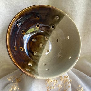 May include: A round ceramic berry bowl featuring a two-tone glaze with a glossy dark brown finish on one side and a speckled cream finish on the other. The bowl is perforated with numerous small circular drainage holes throughout its surface.