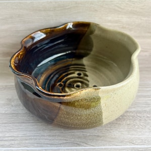 May include: A round ceramic soap dish with a wavy, scalloped rim. The interior features a dark brown and black glossy glaze with a series of drainage holes at the bottom. The exterior has a speckled beige finish that transitions into a dark brown.