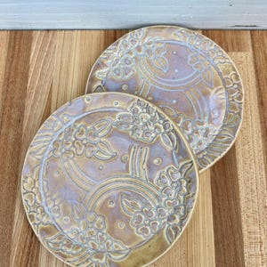 Op de afbeelding: Twee ronde keramische borden met een glanzende afwerking in een mix van lavendel en goudbruine tinten. Het oppervlak van elk bord is versierd met reliëf bloemmotieven en gebogen lijnen, wat zorgt voor een textuur over het hele oppervlak.