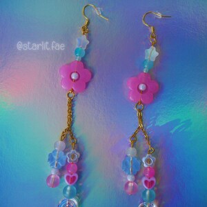 May include: A pair of dangle earrings with a floral theme. Each earring features a pink flower, gold chain, and various beads in shades of pink, blue, and clear. The text "@starlit.fae" is visible.