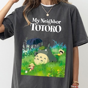 May include: Dark gray t-shirt featuring the text "My Neighbor Totoro" above a colorful graphic of Totoro and other characters in a grassy field. The design includes a large, gray Totoro character and two smaller figures.
