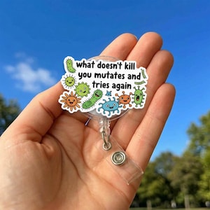 Pode incluir: Um porta-crachá retrátil branco e transparente com um desenho de desenho animado de vários micróbios e o texto "what doesn't kill you mutates and tries again". O porta-crachá tem uma alça de plástico transparente e um clipe de metal.
