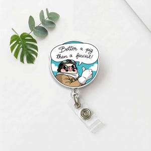 May include: A round, white badge reel featuring a cartoon pig in a pilot's outfit giving a thumbs up. The pig is wearing goggles and a scarf. The speech bubble says, "Better a pig than a fascist!" The reel has a clear plastic strap.