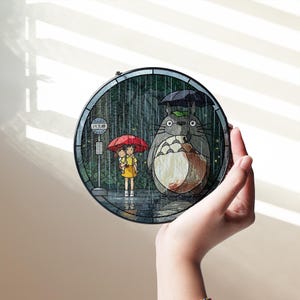 Puede incluir: Un panel redondo de vidriera que representa una escena de la pel&iacute;cula de animaci&oacute;n "Mi vecino Totoro". La obra de arte muestra a Totoro con un paraguas y dos ni&ntilde;os en una parada de autob&uacute;s bajo la lluvia. El panel est&aacute; sostenido por una mano.