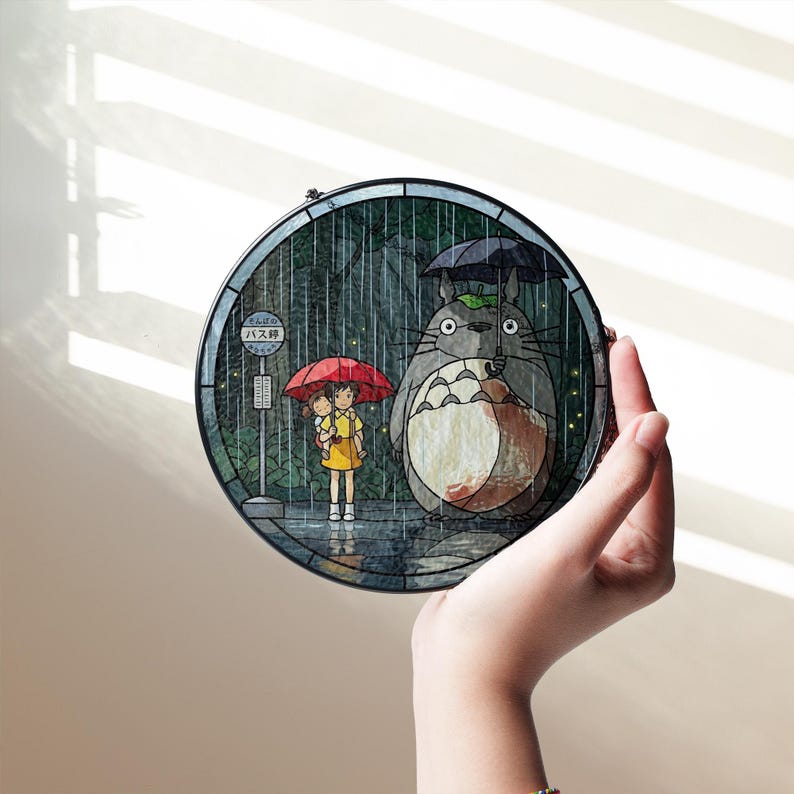 Puede incluir: Un panel redondo de vidriera que representa una escena de la pel&iacute;cula de animaci&oacute;n "Mi vecino Totoro". La obra de arte muestra a Totoro con un paraguas y dos ni&ntilde;os en una parada de autob&uacute;s bajo la lluvia. El panel est&aacute; sostenido por una mano.
