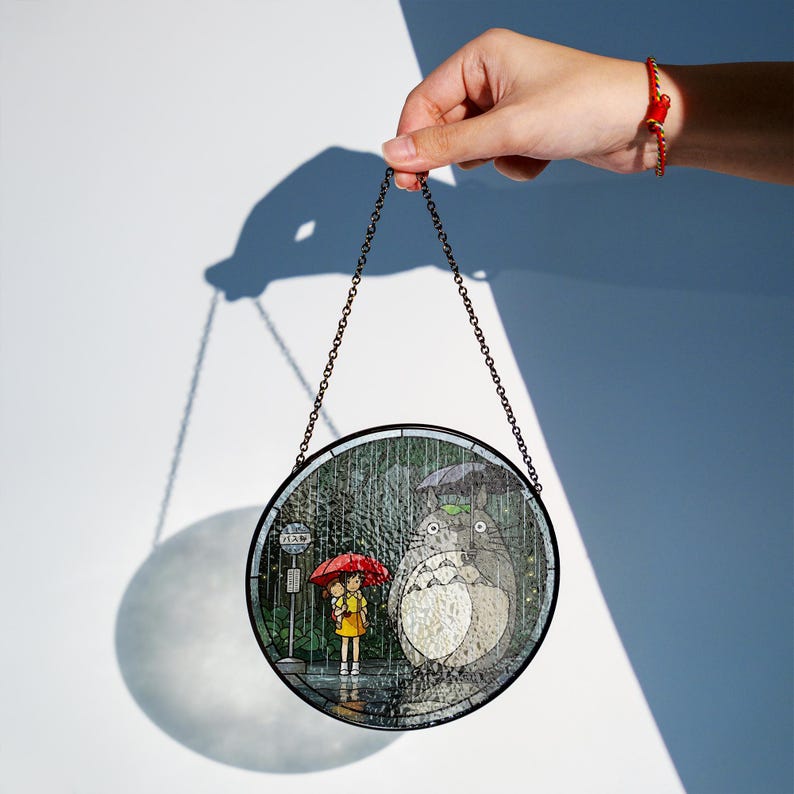 Puede incluir: Panel de vidriera redondo que representa una escena de la pel&iacute;cula de animaci&oacute;n "Mi vecino Totoro". La obra de arte representa un d&iacute;a lluvioso con una ni&ntilde;a y Totoro bajo paraguas. El panel est&aacute; suspendido por una cadena.