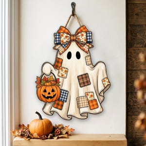 May include: A hanging wall decoration shaped like a ghost, featuring a cream-colored body with various plaid and floral fabric patches. The ghost holds an orange jack-o'-lantern bucket filled with autumn leaves and is topped with a large plaid bow.