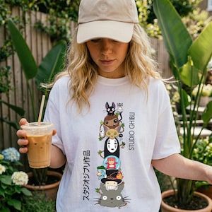 May include: White t-shirt featuring a stacked graphic of Studio Ghibli characters, including Totoro, No-Face, and others. The design includes Japanese text and the words "STUDIO GHIBLI" in black. The person is wearing a beige baseball cap.