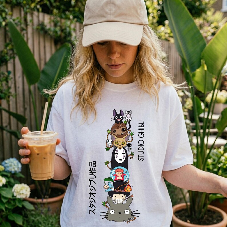 May include: White t-shirt featuring a stacked graphic of Studio Ghibli characters, including Totoro, No-Face, and others. The design includes Japanese text and the words "STUDIO GHIBLI" in black. The person is wearing a beige baseball cap.