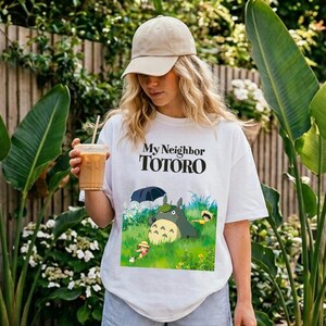 May include: White t-shirt featuring the text "My Neighbor Totoro" above a colorful graphic of the animated characters Totoro and friends in a grassy field. The shirt is made of a soft material.