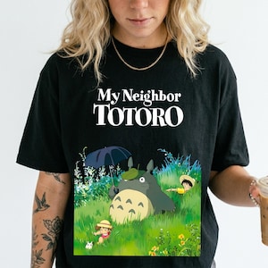 May include: Black t-shirt featuring the text "My Neighbor Totoro" in white. The graphic depicts Totoro with an umbrella, a child, and a cat in a grassy field. The shirt is a crew neck style.