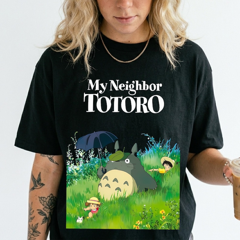May include: Black t-shirt featuring the text "My Neighbor Totoro" in white. The graphic depicts Totoro with an umbrella, a child, and a cat in a grassy field. The shirt is a crew neck style.