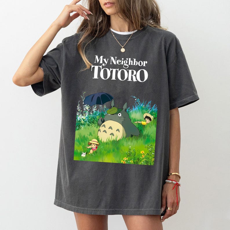 May include: Dark gray t-shirt featuring the text "My Neighbor Totoro" above a colorful graphic of Totoro and other characters in a grassy field. The design includes a large, gray Totoro character and two smaller figures.