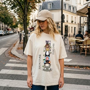 May include: Cream-colored t-shirt featuring a stacked graphic of Studio Ghibli characters, with Japanese text and the words "STUDIO GHIBLI" in black. The shirt is worn by a person wearing a beige cap and blue jeans.