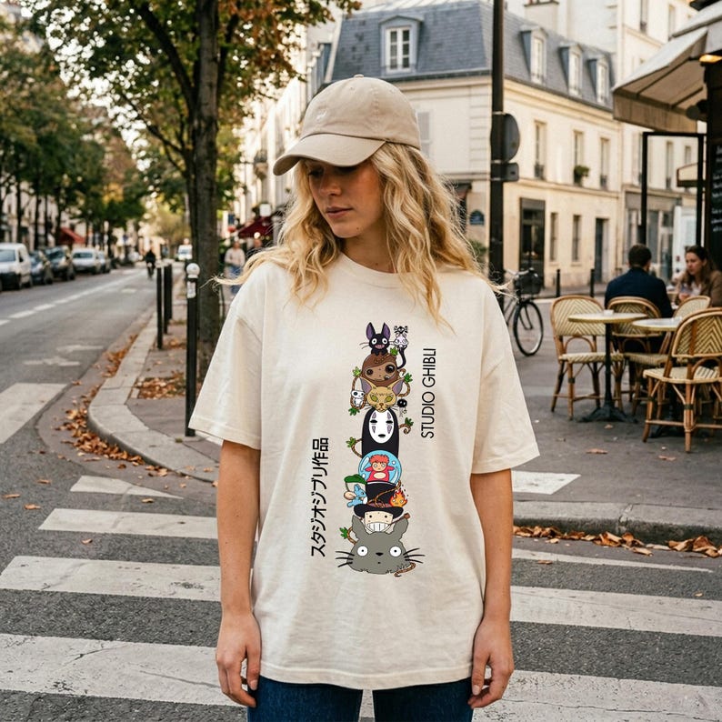 May include: Cream-colored t-shirt featuring a stacked graphic of Studio Ghibli characters, with Japanese text and the words "STUDIO GHIBLI" in black. The shirt is worn by a person wearing a beige cap and blue jeans.