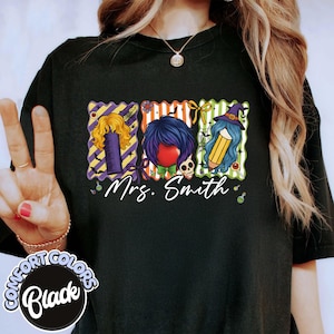 Op de afbeelding: Zwart Comfort Colors T-shirt met een afbeelding van drie gestileerde haar- en accessoire-ontwerpen. De tekst "Mrs. Smith" staat eronder. Het shirt heeft een ronde hals en korte mouwen.