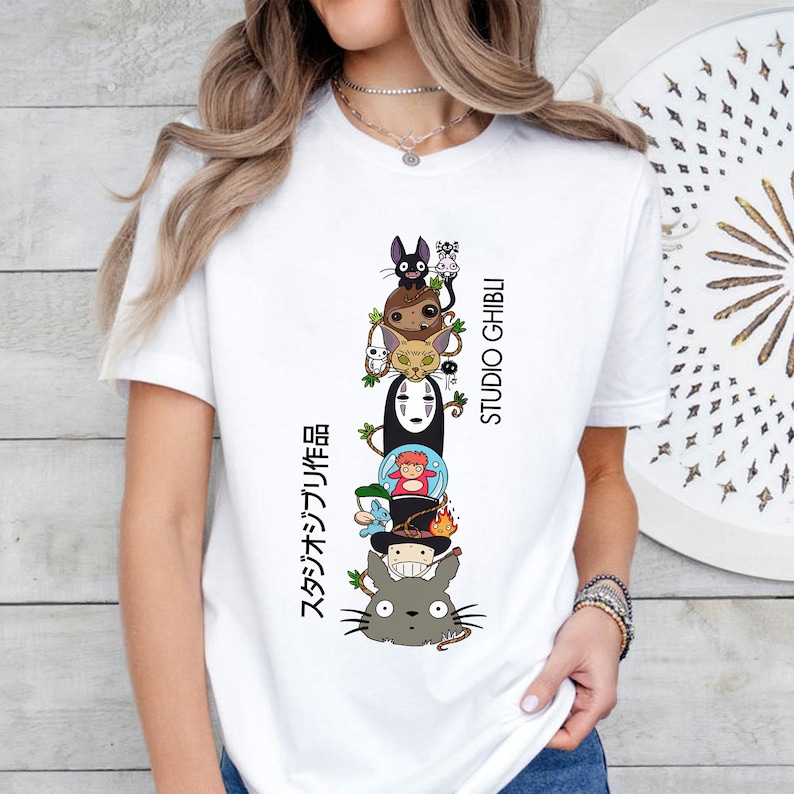 May include: White t-shirt featuring a stacked graphic of Studio Ghibli characters, including Totoro, No-Face, and others. The text "STUDIO GHIBLI" and Japanese text are printed vertically on the right and left sides of the graphic.