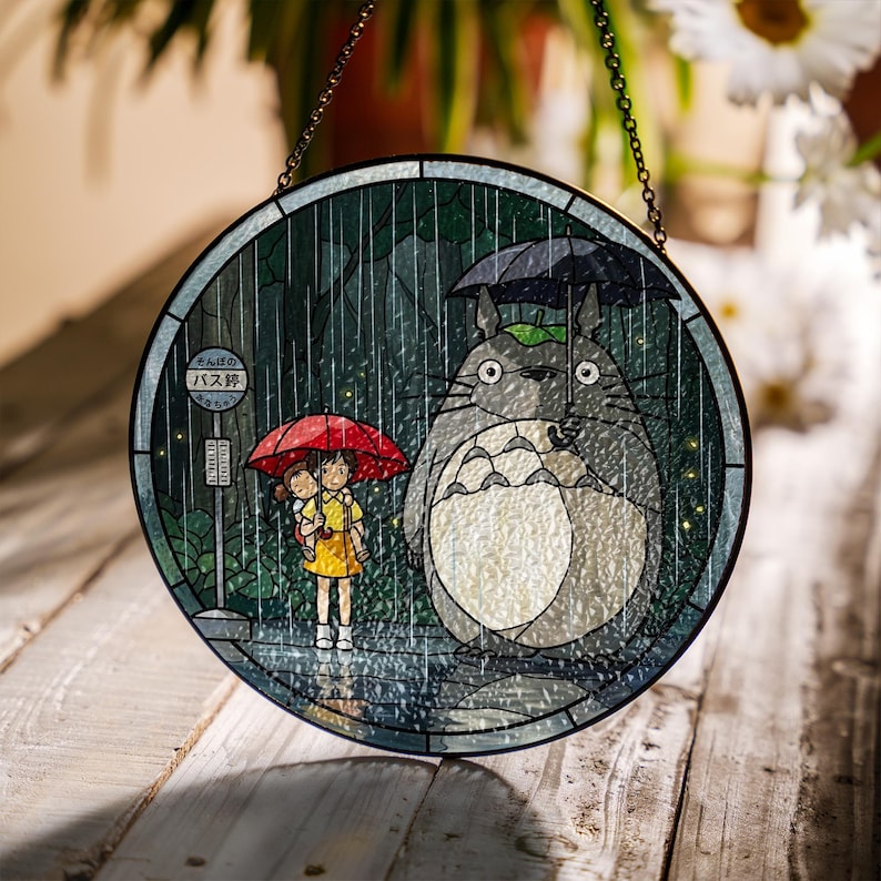 Puede incluir: Panel de vidriera redondo que representa una escena de la pel&iacute;cula de animaci&oacute;n Mi vecino Totoro. La obra de arte muestra a Totoro con un paraguas, dos ni&ntilde;os bajo un paraguas rojo y una parada de autob&uacute;s bajo la lluvia. El panel tiene un borde de metal negro y una cadena para colgar.