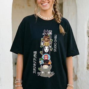 May include: Black t-shirt featuring a stacked graphic of Studio Ghibli characters, including Totoro and No-Face. The design includes Japanese text and the words "STUDIO GHIBLI" in white. The shirt has short sleeves and a relaxed fit.