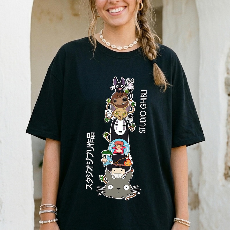 May include: Black t-shirt featuring a stacked graphic of Studio Ghibli characters, including Totoro and No-Face. The design includes Japanese text and the words "STUDIO GHIBLI" in white. The shirt has short sleeves and a relaxed fit.