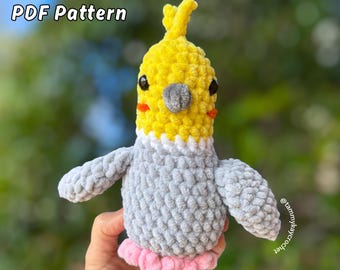 Op de afbeelding: Een gehaakte Lil Cockatiel-knuffel in geel, grijs en roze. De vogel heeft zwarte ogen, oranje wangen en een grijze snavel. De tekst "NO SEW PDF Pattern" en "Lil Cockatiel" zijn zichtbaar.