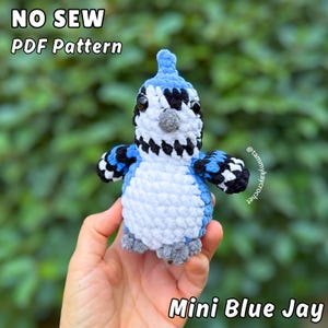 May include: A handmade, mini blue jay plushie with black, white, and blue accents. The bird has a textured, crocheted appearance and is held in a person's hand. Text on the image reads "NO SEW PDF Pattern" and "Mini Blue Jay".