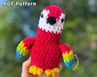 Op de afbeelding: Een handgemaakte gehaakte papegaai in rood, wit, zwart, geel, blauw en groen. De tekst "NO SEW PDF Pattern" en "Lil Parrot Macaw" worden weergegeven. De papegaai heeft een getextureerd, zacht uiterlijk.