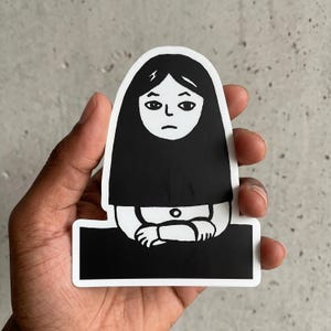May include: A die-cut vinyl sticker featuring a black and white illustration of a person with long dark hair, a neutral facial expression, and arms crossed, positioned behind a solid black rectangular base with a white border outline.