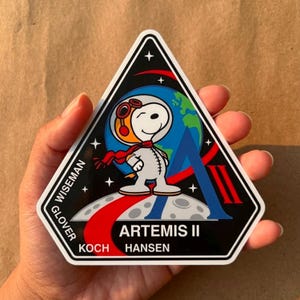 May include: A triangular Artemis II mission patch featuring Snoopy in a spacesuit, with a red scarf and goggles. The patch includes the Earth, stars, and the names of the crew: Wiseman, Glover, Koch, and Hansen.