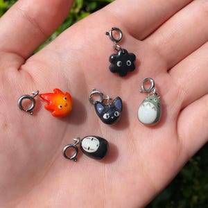May include: A collection of colorful anime-inspired charms. The charms include a black soot sprite, a fiery orange flame, a black cat, a gray and white creature, and a masked figure. Each charm has a silver clasp.