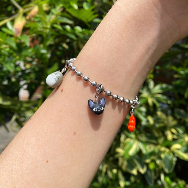 May include: A silver-colored charm bracelet with three dangling charms. The charms include a gray cat, a black cat, and a red pepper. The bracelet is on a wrist with a green leafy background.