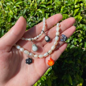 May include: Three pearl necklaces with various charms. The charms include a Totoro figure, a black soot sprite, a black cat, a small orange fire demon, and a character from Spirited Away. The necklaces are held in a hand.