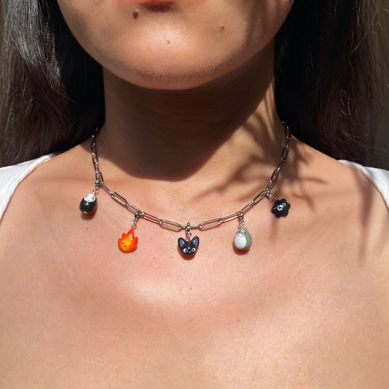 May include: A silver-toned chain necklace featuring six colorful charms. The charms include a black soot sprite, a fiery orange flame, a black cat, a gray Totoro, a black flower, and a white and black charm.