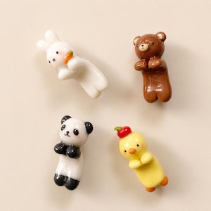 Pode incluir: Um conjunto de quatro suportes para pauzinhos de cerâmica em forma de animais. A coleção inclui um coelho branco com uma cenoura laranja, um urso castanho, um panda preto e branco e um pintainho amarelo com uma pequena maçã vermelha na cabeça.