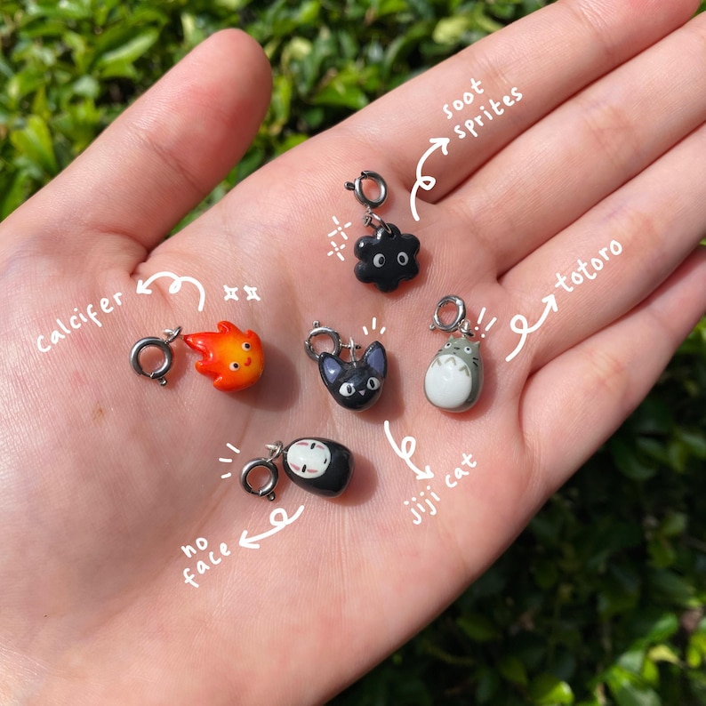 May include: A collection of small, colorful charms featuring characters from animated films. The charms include a black soot sprite, a red and orange Calcifer, a black Jiji cat, a gray and white Totoro, and a black No-Face.