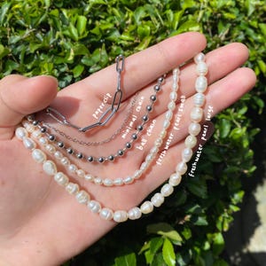 May include: A hand displays a collection of necklaces. The necklaces include a paperclip chain, a minimal chain, a ball chain, and two freshwater pearl necklaces labeled "small" and "large". The chains are silver and the pearls are white.