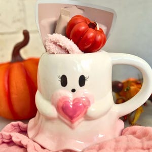 May include: A white ghost-shaped mug with a pink heart, holding a red pumpkin and pink plush. An orange pumpkin sits behind the mug. The mug is on a pink cloth, with fall-themed decor.