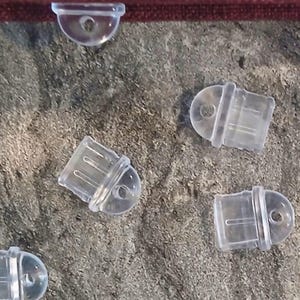 May include: A selection of small, clear plastic balloon clips scattered on a textured grey surface. Each clip features a rounded top with a hole and a rectangular base with vertical grooves. These clips are designed to secure balloons.