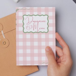 May include: A person holding a pink and white gingham card with the words "Dopamine Menu" in a green frame. A brown envelope and a white pen are also visible on a gray surface.