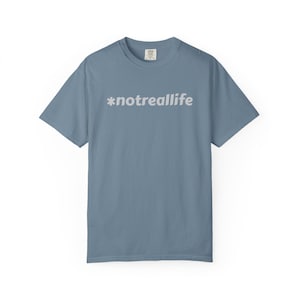 May include: A dusty blue short-sleeved t-shirt with the text "*notreallife" printed in light gray across the chest. The shirt has a crew neck and a relaxed fit. The fabric appears to be soft and comfortable.
